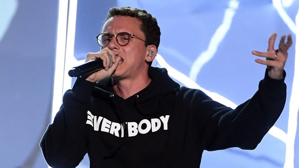 The rapper is performing on stage, wearing round glasses and a black hoodie with the text "EVERYBODY" printed in white, while holding a microphone in one hand and gesturing with the other.