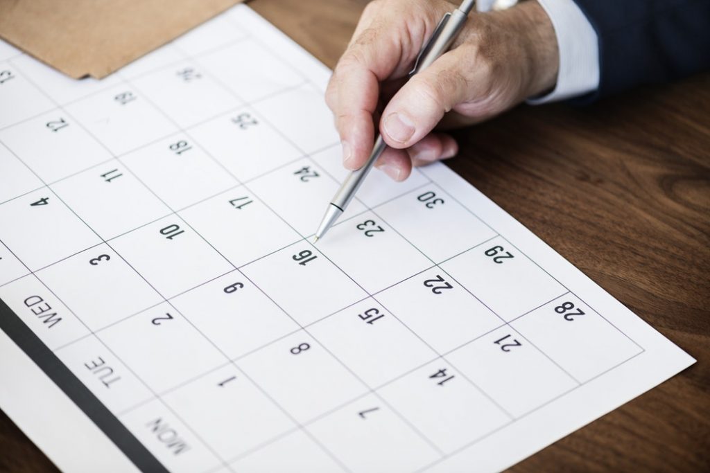 A person in a suit holding a silver pen, poised to write on a paper calendar laid out on a wooden desk, highlighting the concept of monthly planning.