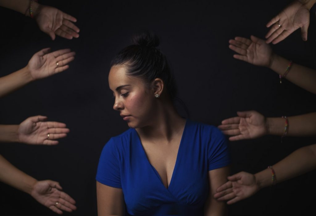 A woman in a blue shirt sits with her head down, looking contemplative and slightly distressed, surrounded by numerous hands reaching toward her from all directions against a dark background.