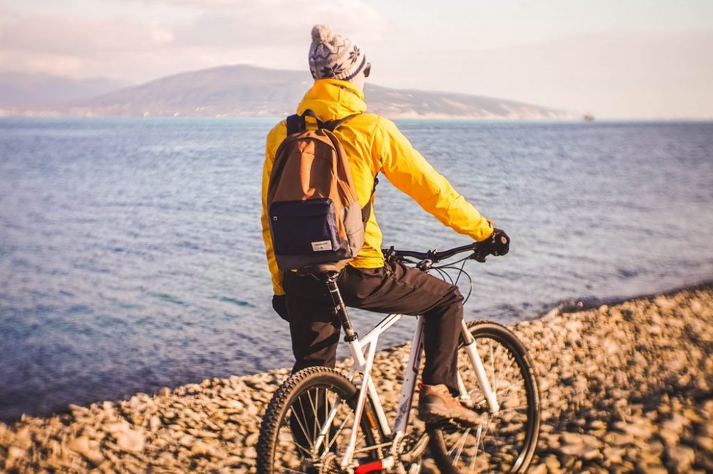 A person wearing a bright yellow jacket, a knit hat, and a backpack, riding a mountain bike along a rocky lakeshore path with a calm body of water in the background under a soft, golden light.