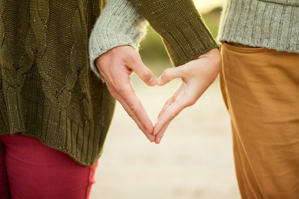 A close-up of two people standing together during an autumn walk; their hands are positioned together to form a heart shape against the backdrop of their clothing, with the soft-focus fall landscape visible in the background.