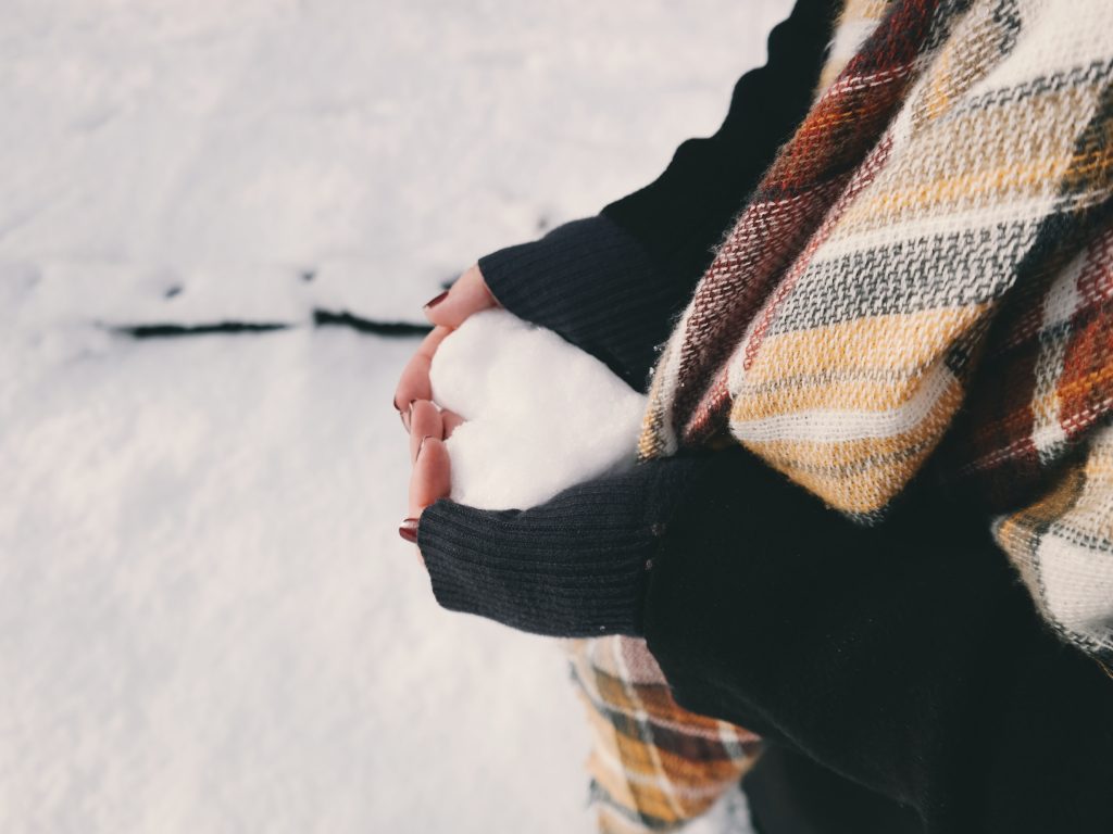 A close-up, side-view shot of a person wearing a black sweater and a brown and white plaid scarf, holding a heart-shaped mound of snow in their cupped hands against a snowy, white background.