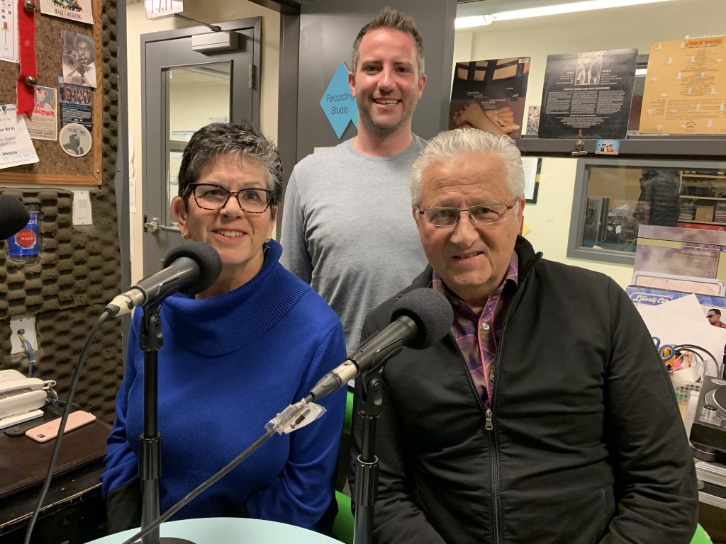 A man and a woman in a radio studio setting, both smiling and speaking into microphones. The woman is wearing a bright blue sweater, and the man beside her is wearing a dark, zip-up jacket over a plaid shirt. Behind them stands another man, also smiling, in a light-coloured grey sweater. The background includes studio equipment and shelves with various items.