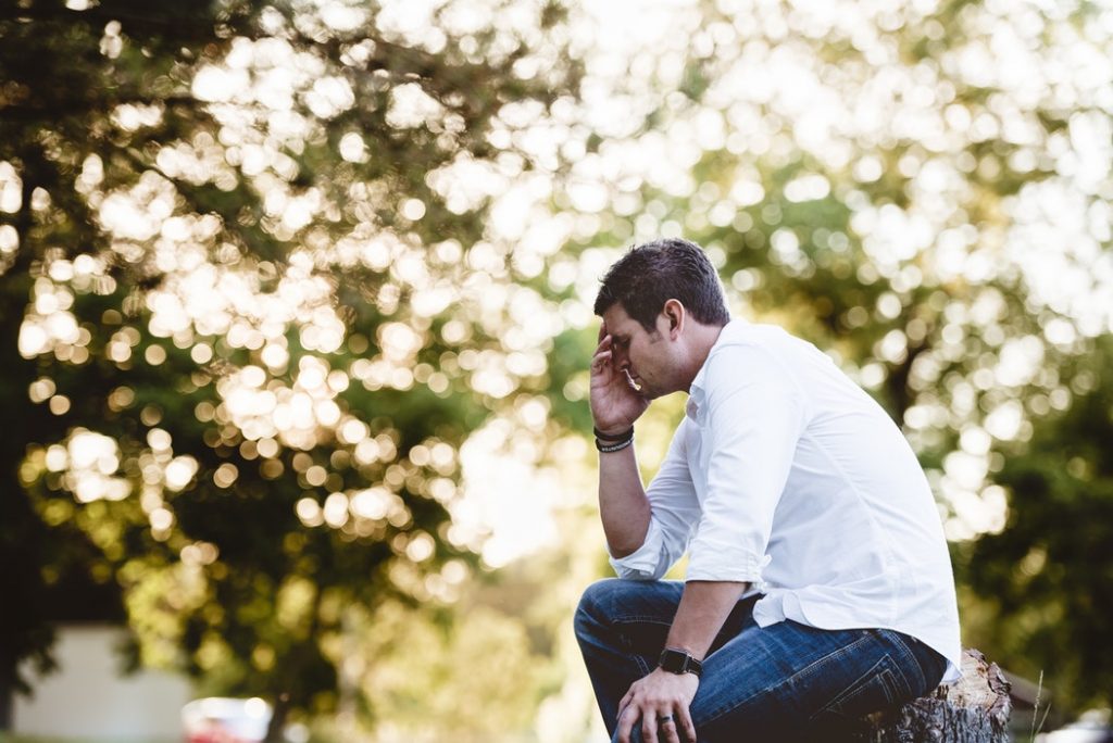A young man sitting on a tree stump in a park, leaning forward with his head in his hands, appearing overwhelmed or distressed. The background shows trees with dappled sunlight filtering through the leaves, creating a soft, warm atmosphere.