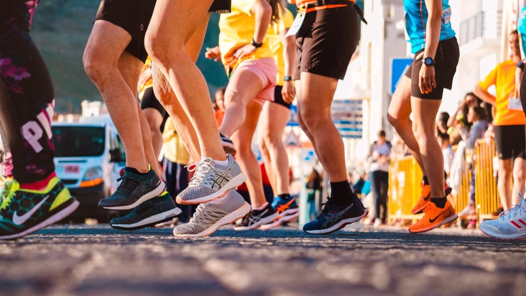 A close-up, low-angle shot of the legs and colourful running shoes of a crowded pack of marathon participants actively running on a paved city street during a race.