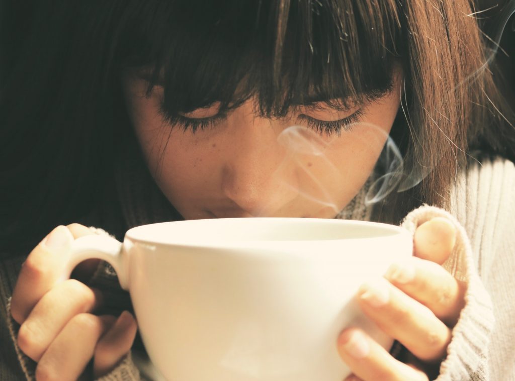 A close-up of a young woman with her eyes closed, tenderly holding a warm white mug in both hands. Steam rises from the coffee, creating a cozy and serene atmosphere as she enjoys the warmth on a cool day.