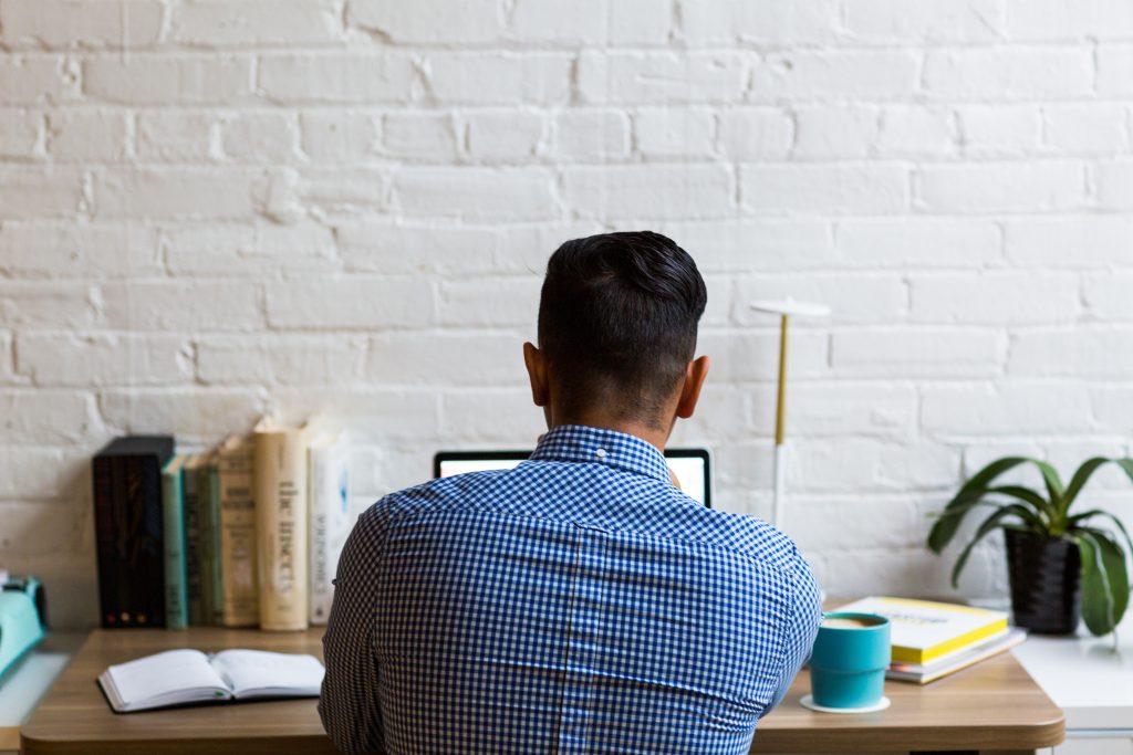 A person seen from behind, wearing a blue plaid shirt, sitting at a wooden desk and working on a computer. The desk is organized with a small plant, a stack of books, and a notebook, set against a white brick wall.