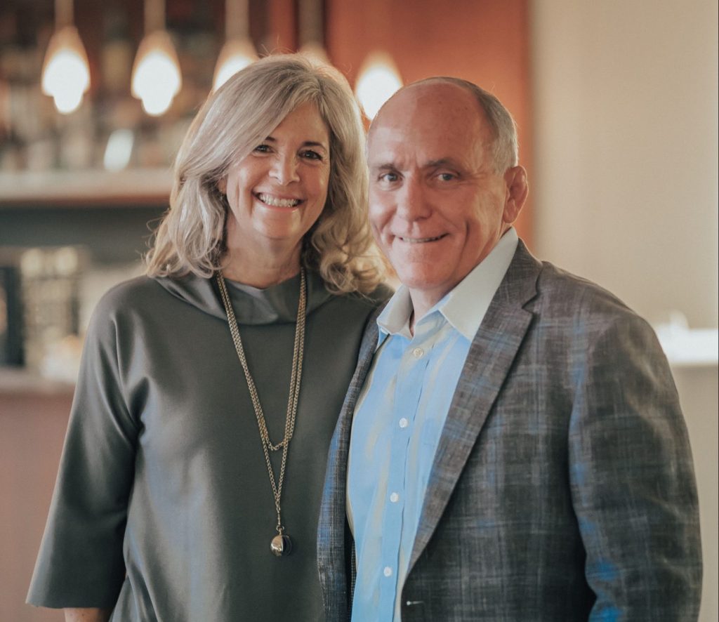 A portrait of Ellen Novack and John Park, a woman with blonde hair wearing a grey top and long necklace standing beside a man with short hair wearing a blue shirt and a patterned blazer, both smiling warmly at the camera.