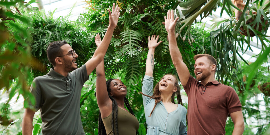 Four people smiling and raising their hands together in a high-five gesture inside a lush green greenhouse.