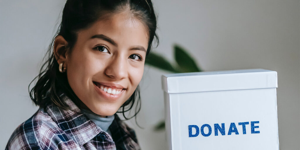 A close-up photograph of a smiling young woman with dark hair tied back, wearing a plaid shirt. She is standing next to a white donation box that has the word "DONATE" written across it in blue capital letters.