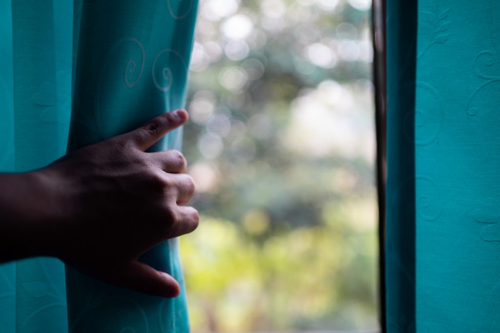 A hand reaches out to open a teal-colored curtain, revealing a soft-focus, sunlit view of the outdoors through a window. The image suggests a sense of transition, hope, or new beginnings.