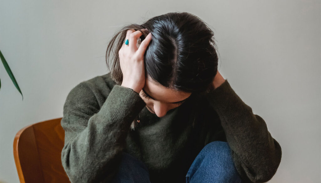 A close-up photograph of a young woman sitting down and looking downward with a distressed expression. She has dark hair and is holding both hands tightly to her head in a gesture of overwhelm or pain, wearing a dark green sweater and jeans.
