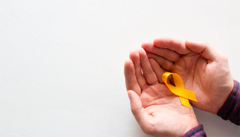 A close-up photograph of two cupped hands gently holding a folded yellow awareness ribbon against a plain, off-white background.