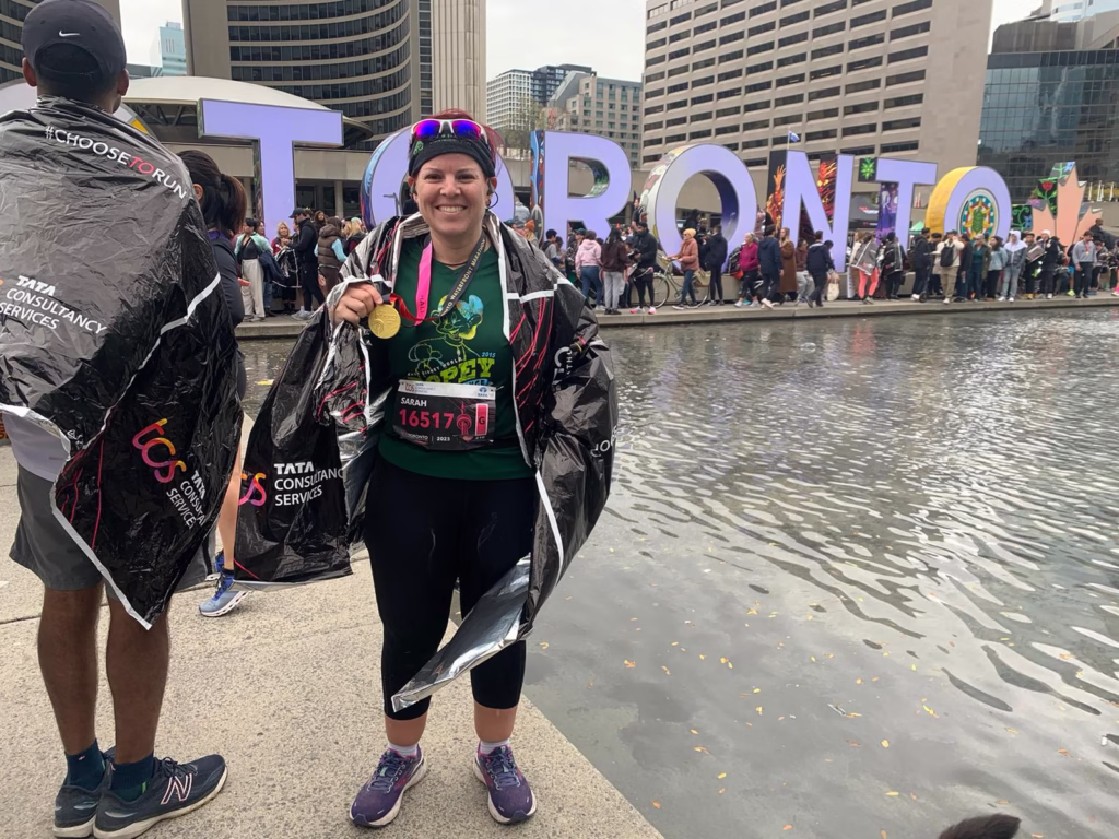 A cheerful, full-length outdoor portrait of Sarah Swartz smiling broadly at Nathan Phillips Square in Toronto. She is wearing black running tights, a green race t-shirt with her bib number 16517, and has a finisher's medal hanging around her neck. She is wrapped in a black plastic "Tata Consultancy Services" thermal race blanket, with the vibrant, large multi-coloured "TORONTO" sign and a crowd of event participants visible in the background by the water.