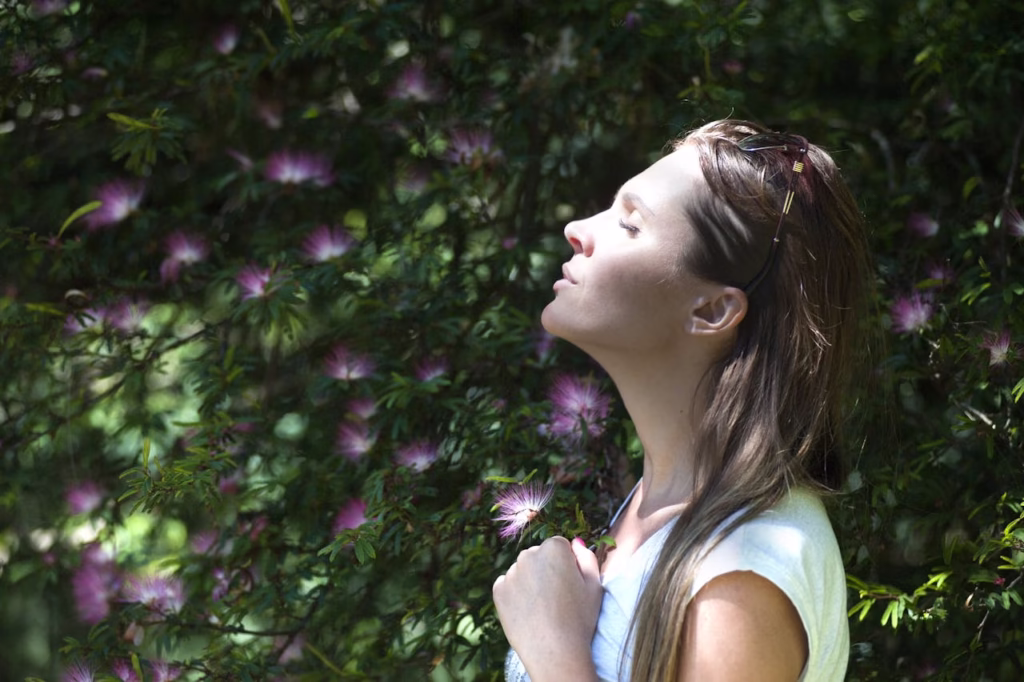 A close-up profile shot of a young woman with long brown hair, closed eyes, and a peaceful expression, tilting her head back to breathe deeply in the sunlight. She is wearing a white top and is surrounded by lush green foliage filled with pink silk tree blossoms in soft focus.
