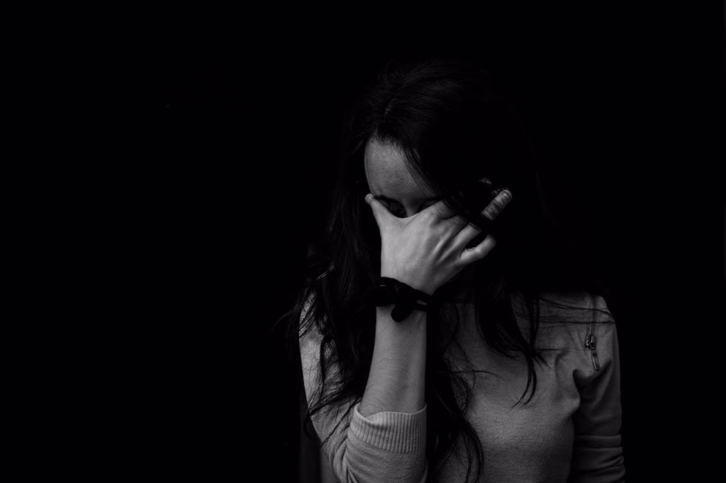 A high-contrast, black-and-white close-up shot of a young woman experiencing deep distress against a dark background. She has long, dark hair and is holding one hand up to cover her face, with her fingers pressed tightly against her forehead and eyes in an expression of overwhelm or grief.
