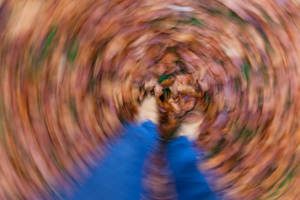 A motion-blurred, first-person perspective shot looking down at blue sleeves and gloved hands reaching into a swirling vortex of autumn leaves.