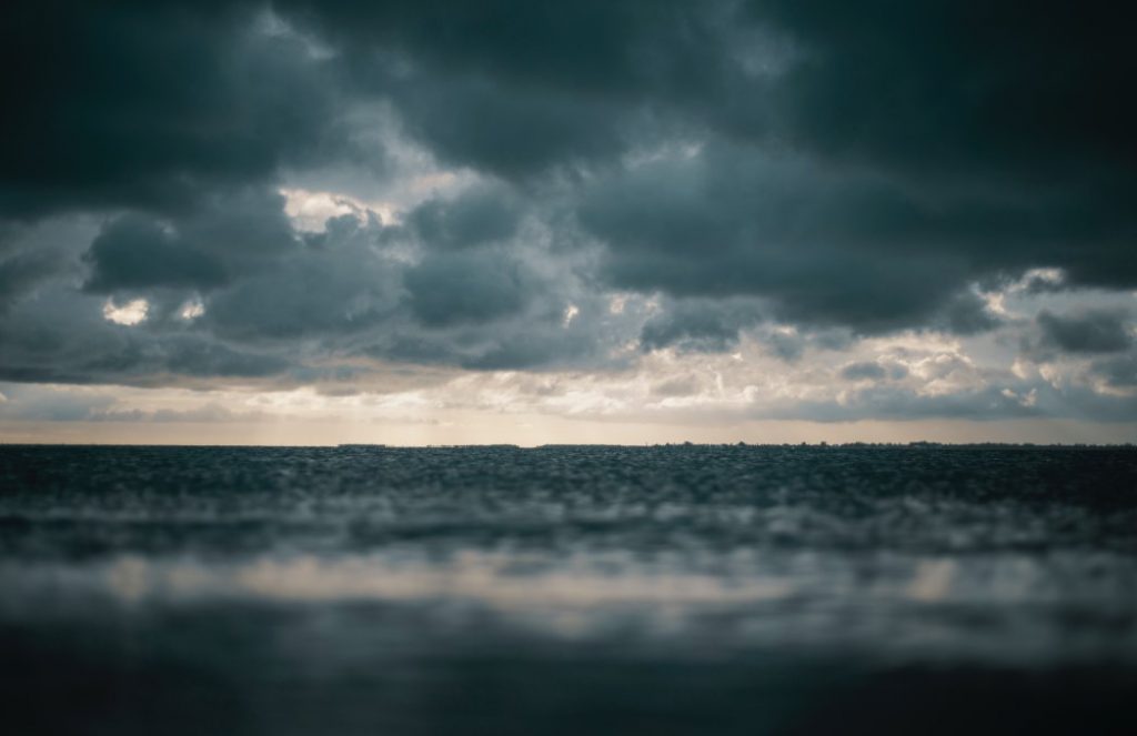 A panoramic view of a vast, calm body of water meeting a dark, stormy sky filled with heavy, grey clouds, with a sliver of light breaking through on the horizon.