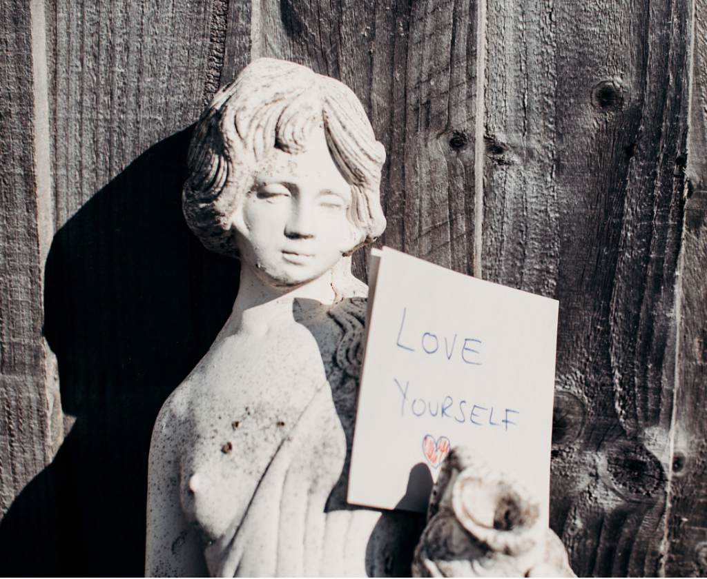 A close-up of a weathered stone bust of a woman's head and shoulders placed in front of a rustic, dark-stained wooden plank wall. The woman is holding a piece of paper that says "LOVE YOURSELF" in handwritten letters, with a small, colorful heart drawing at the bottom.
