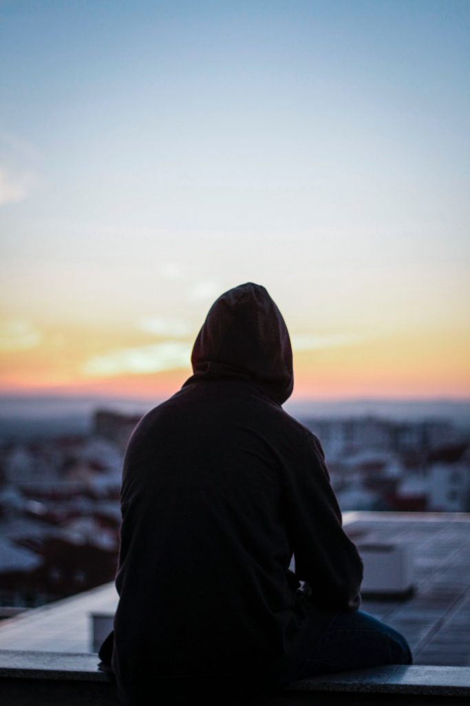 A silhouette of a person wearing a hoodie, sitting and looking out over a city at sunset, symbolizing reflection and the journey toward mental wellness offered at Eli's Place.