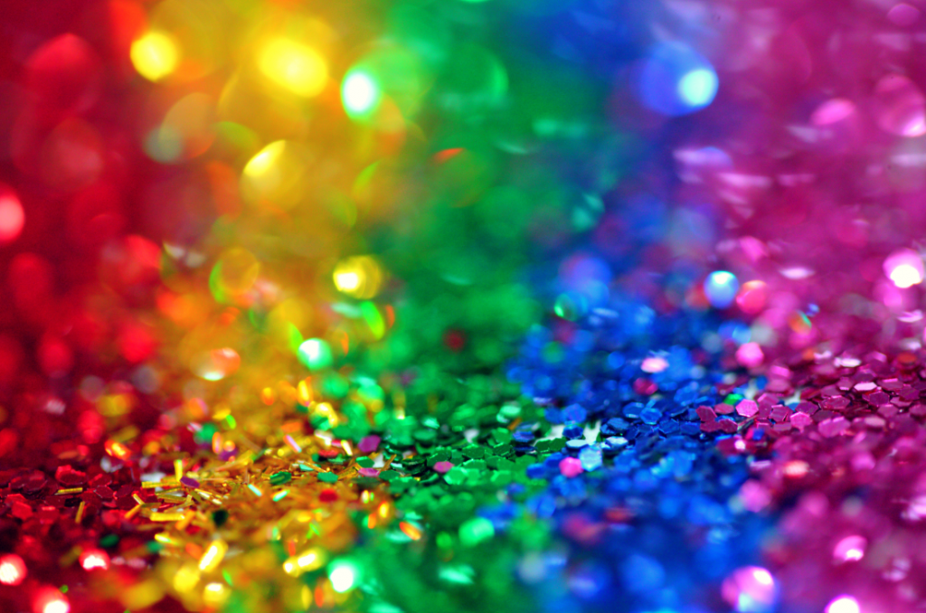 A vibrant, out-of-focus background filled with sparkling, multi-colored glitter transitioning through the spectrum of the rainbow, from warm reds and yellows on the left to cool greens and blues on the right.