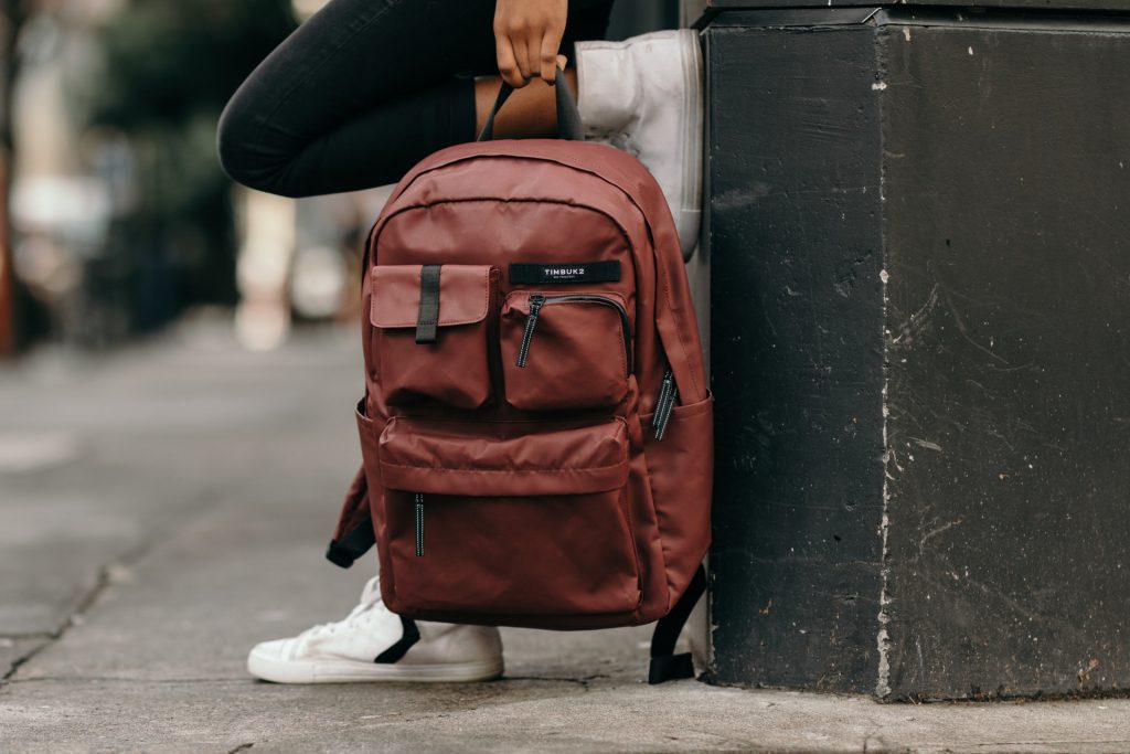 A person sitting on a concrete surface, wearing black jeans and white sneakers, with a dark red Timbuk2 backpack resting beside them. The shot is captured from the waist down, focusing on the backpack in an urban setting.