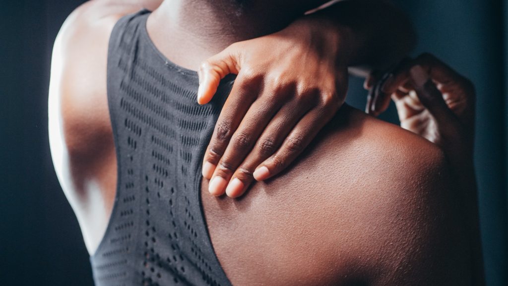 A close-up, over-the-shoulder view of a person wearing a black athletic top, reaching their right hand across to touch or massage their left shoulder blade, which is slightly tense. The lighting is soft and moody, emphasizing the person's back and shoulder.