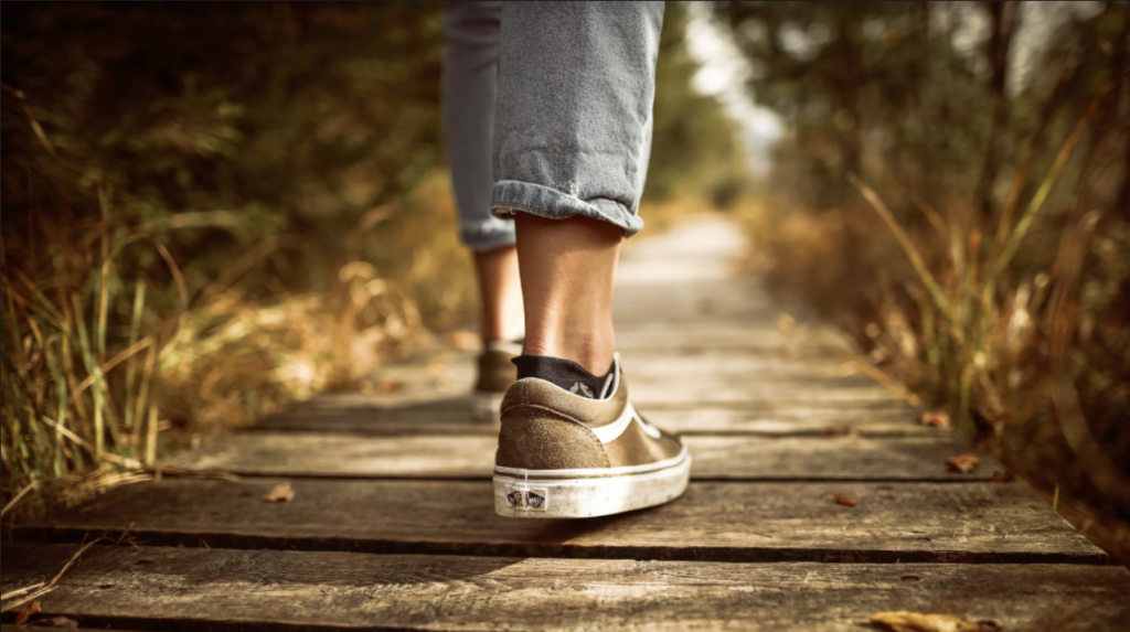 A panoramic crop showing someone's feet and the lower part of their grey pants walking along a path or trail in a sun-dappled woods.