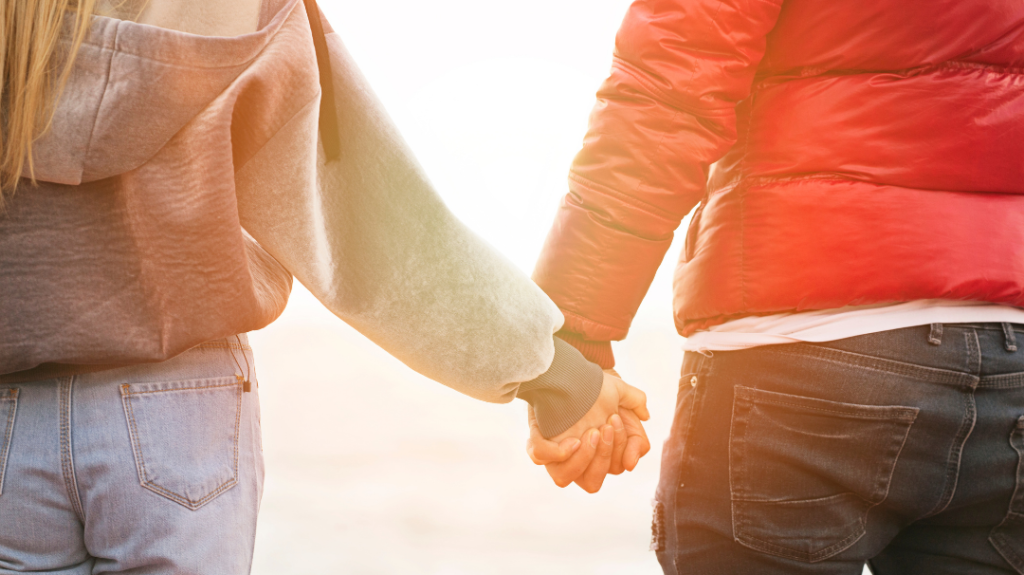A view from behind of two people holding hands while walking outdoors. The person on the left is wearing a light grey hooded sweatshirt and denim jeans, and the person on the right is wearing a bright red winter jacket and dark jeans.