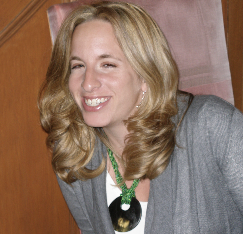 A portrait of a woman with blonde hair smiling warmly at the camera. She is wearing a grey blazer, a green beaded necklace with a large black circular pendant, and delicate earrings.