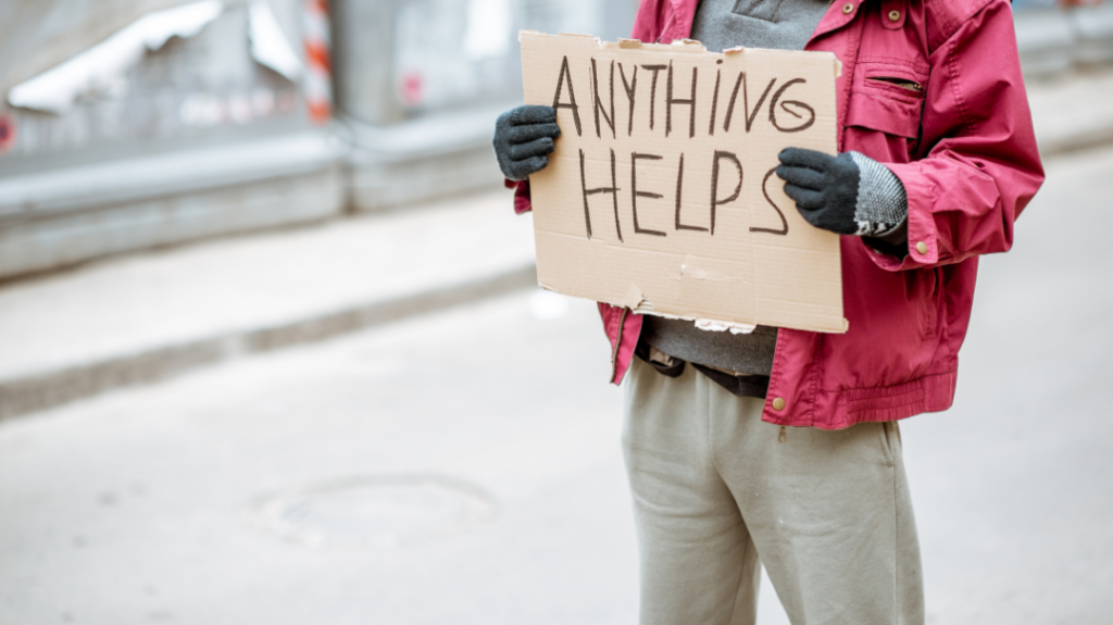 A person wearing a bright red winter jacket and black gloves stands outdoors, holding up a cardboard sign that reads "ANYTHING HELPS" written in black marker.
