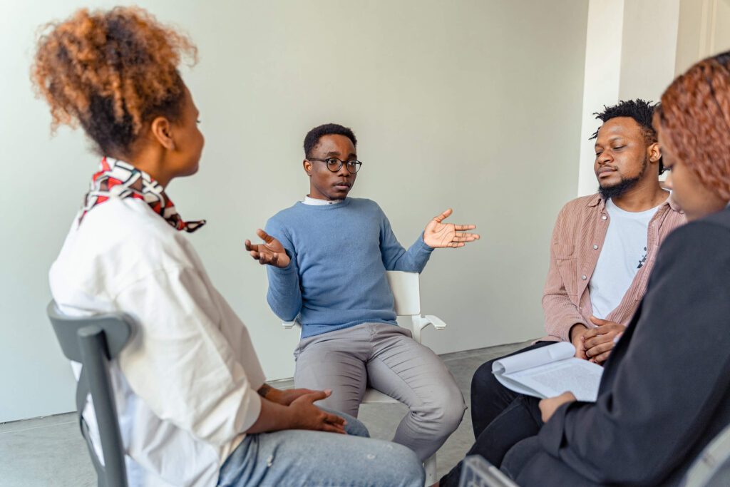 A medium shot of a diverse therapy or support group session indoors. In the center, a young Black man wearing glasses and a light blue sweater sits on a white chair, gesturing with open hands as he speaks. To his right, another Black man in a pink button-down shirt over a white t-shirt listens intently with his hands clasped. On the left and right foreground, two other group members are seen from behind or in profile, one holding a pen and notebook, creating an intimate, collaborative circle.