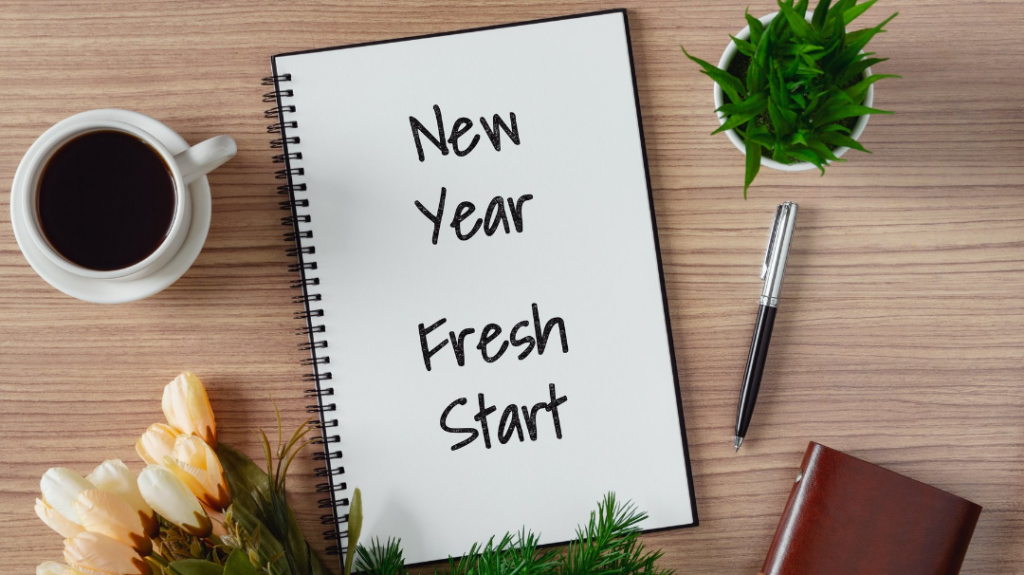 A flat lay arrangement on a wooden desk. On the left is a white mug filled with black coffee and a small bouquet of white tulips. In the center is an open spiral notebook with "New Year Fresh Start" handwritten on the page next to a silver pen. In the top right is a small green potted plant, and in the bottom right is a brown leather notebook next to a small evergreen branch.