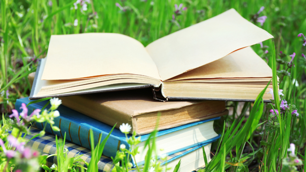 A stack of three hardcover books sitting on green grass surrounded by small purple flowers, with the top book open face-down.