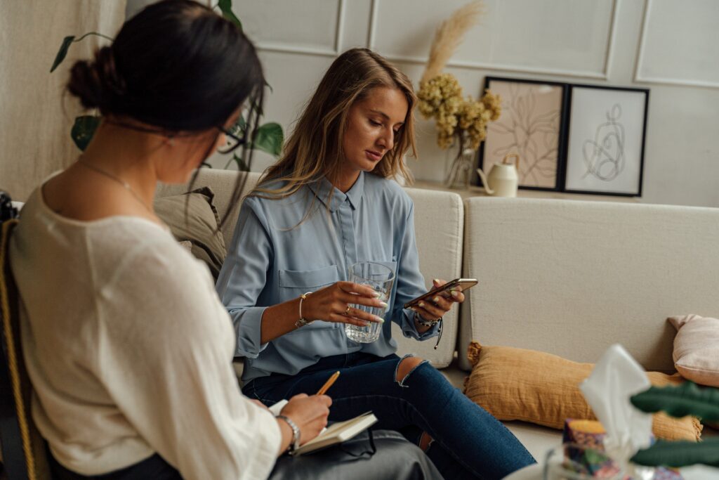 A medium shot of a young woman with long blonde hair and ripped jeans sitting on a white couch indoors. She is looking down at her smartphone while holding a glass of water. Beside her in the foreground, another woman with her dark hair up in a bun is viewed from behind, sitting and taking notes in a notebook.