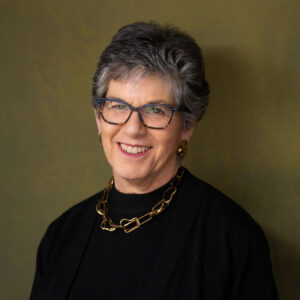 A headshot of a smiling woman with short gray hair and glasses, wearing a black top and a thick gold chain necklace against a textured green background.