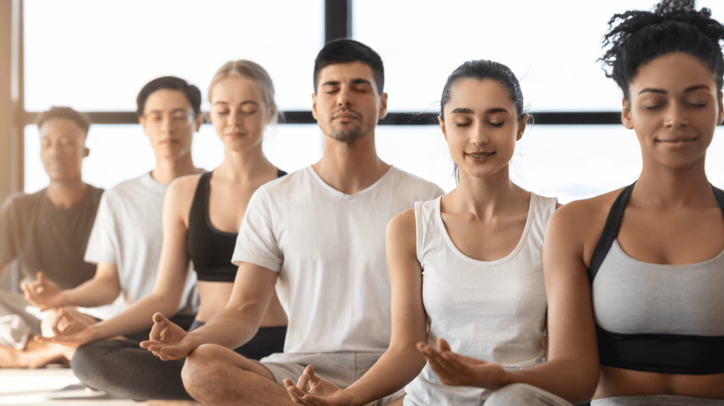 A row of five people sitting cross-legged on yoga mats in a brightly lit studio, meditating with their eyes closed and hands resting on their knees in a mudra position.