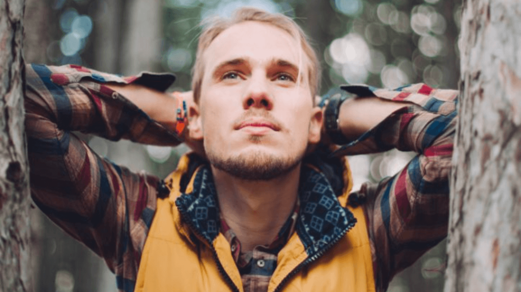 Eli's Place homepage photo: A man looking upwards with his hands resting behind his head, leaning against a tree trunk in a wooded area, wearing a yellow vest over a plaid shirt.
