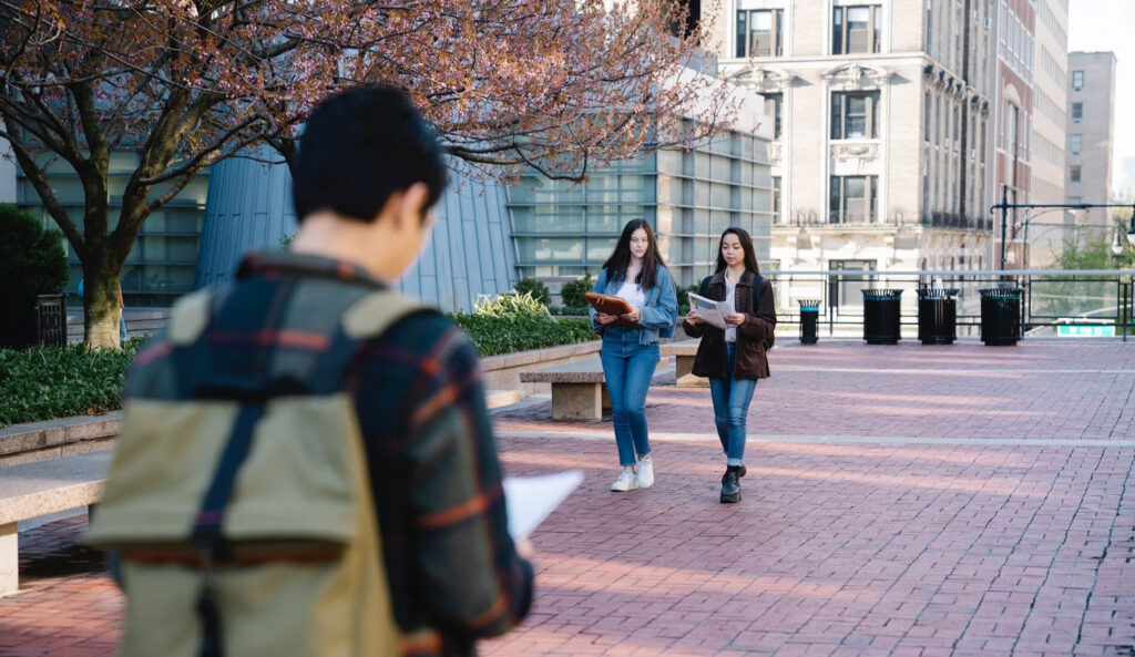 A shallow-focus shot from behind a young person with a backpack, looking toward two young women who are smiling, chatting, and walking together down a wide brick pathway on a school campus or city square.