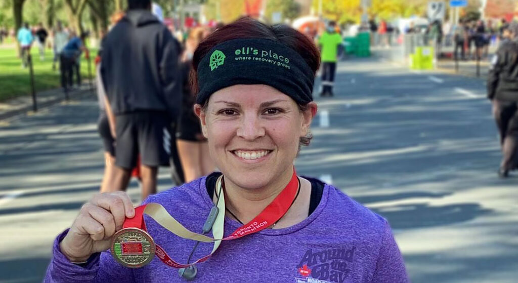 A smiling woman wearing a purple shirt and an "Eli's Place" headband stands outside on a sunny day, proudly holding up a marathon finisher's medal around her neck.