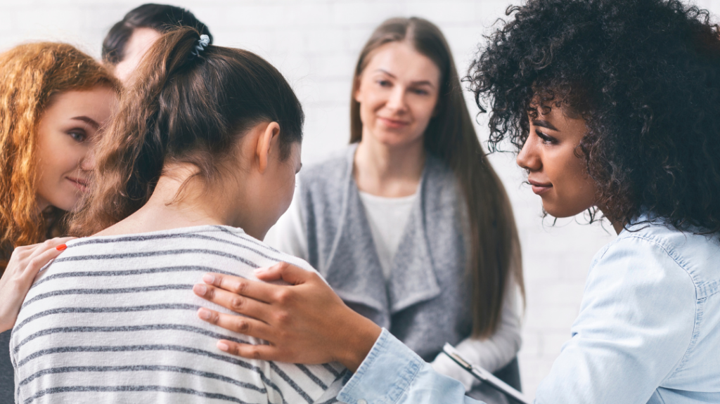 A medium shot of four women standing in a circle, smiling and talking together in a supportive group setting at an Eli's Place event. The woman in the center foreground has her back partially turned, wearing a striped grey and white shirt, while another woman gently places a hand on her shoulder in comfort.