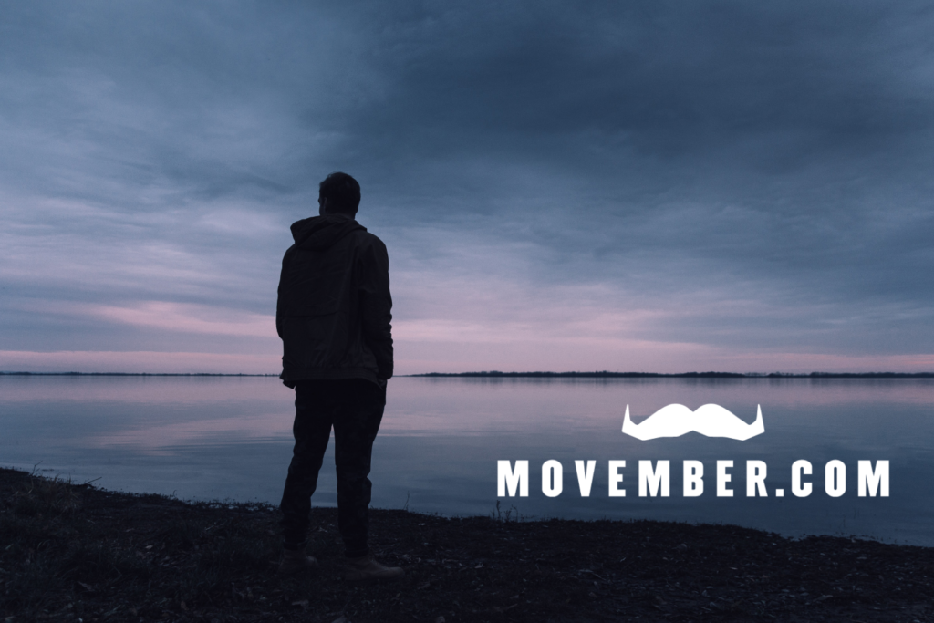 A silhouette of a person standing on a rocky shoreline, looking out over a wide, calm body of water under a dramatic, overcast sky. The white words "MOVEMBER.COM" are centered across the water, topped with a stylized white mustache icon.