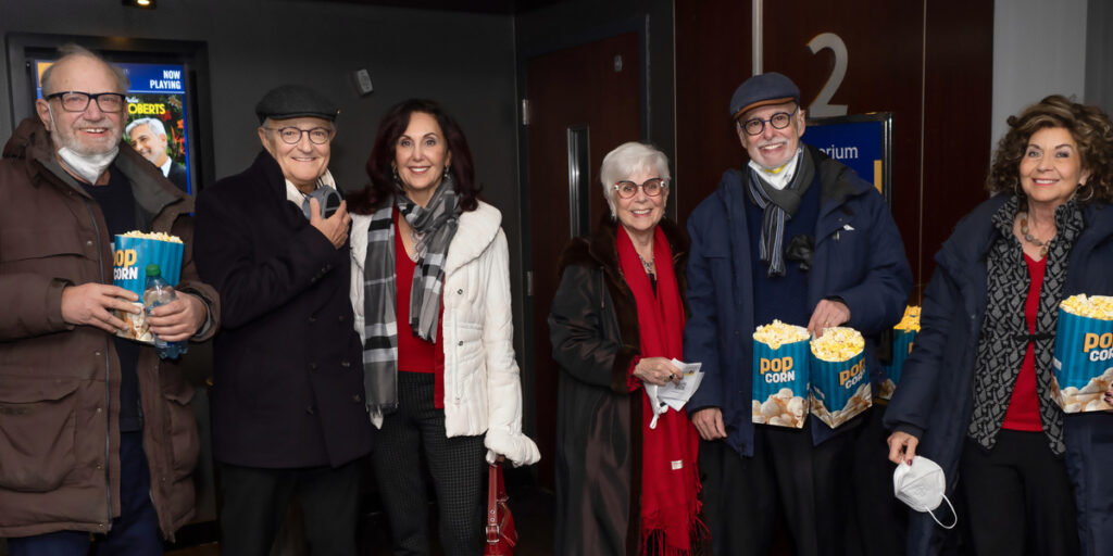 A group of six people standing shoulder-to-shoulder and smiling at an Eli's Place film screening event. They are posed indoors under an "Auditorium 2" sign, wearing winter coats and holding buckets of popcorn.
