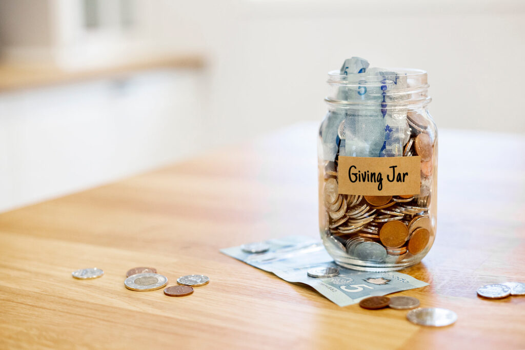 An eye-level close-up shot of a clear glass "Giving Jar" sitting on a smooth light-wood tabletop. The jar is packed with neatly rolled blue five-dollar bills at the top, while the bottom half is filled to the brim with loose coins. A rustic brown paper label is taped to the front of the jar with the words "Giving Jar" handwritten in black marker. A few loose coins and a single blue five-dollar bill lie scattered on the table around the base of the jar, while the background is softly blurred.