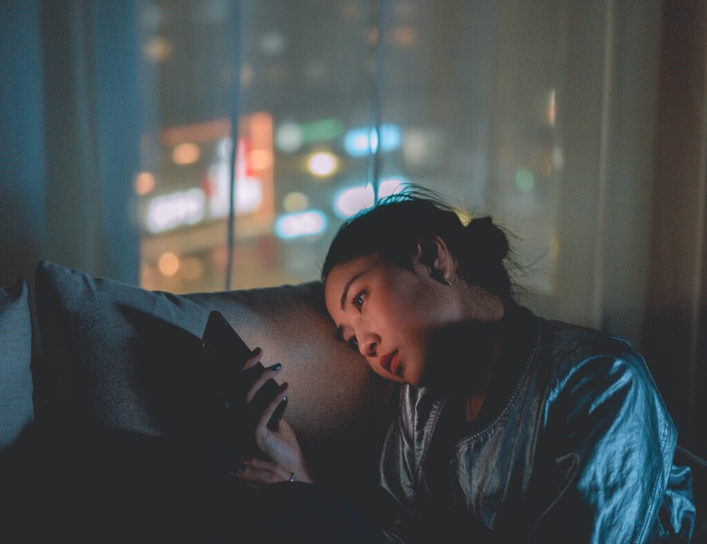 A close-up, dimly lit photograph of a young woman with her hair in a bun, resting her head sideways against a grey couch cushion while looking thoughtfully at her smartphone. The warm, colorful lights of a city skyline at night are softly blurred in the background through a window.