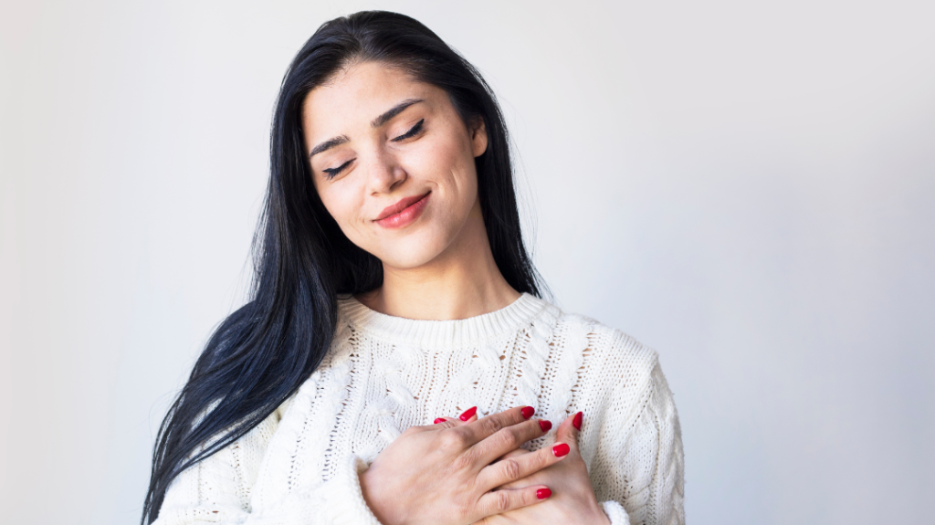 A close-up, front-facing portrait of a young woman with long, straight black hair, smiling gently with her eyes closed. She has fair skin and red nail polish, and her hands are gently crossed over her heart. She is wearing a cozy, cream-colored, textured cable-knit sweater against a plain, light grey background.