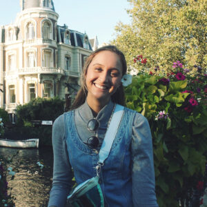 A medium portrait of a smiling young woman with long, straight dark brown hair, wearing a flower in her hair behind her left ear. She is looking directly at the camera with a joyful expression. She wears a grey turtleneck under a denim pinafore dress with a light blue strap from a small bag slung over her shoulder. The background features a beautiful historic European-style white building with intricate architecture, surrounded by lush green trees and vibrant pink flowers.