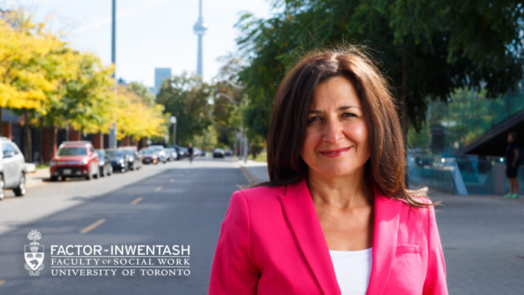 A professional headshot portrait of Dr. Toula Kourgiantakis, a woman with shoulder-length dark brown hair, smiling gently at the camera. She is wearing a bright pink blazer over a white top. The background shows a blurred city street scene with trees showing autumn foliage and the CN Tower visible in the distance. The logo for the "FACTOR-INWENTASH FACULTY OF SOCIAL WORK | UNIVERSITY OF TORONTO" is overlaid in white in the bottom left corner.