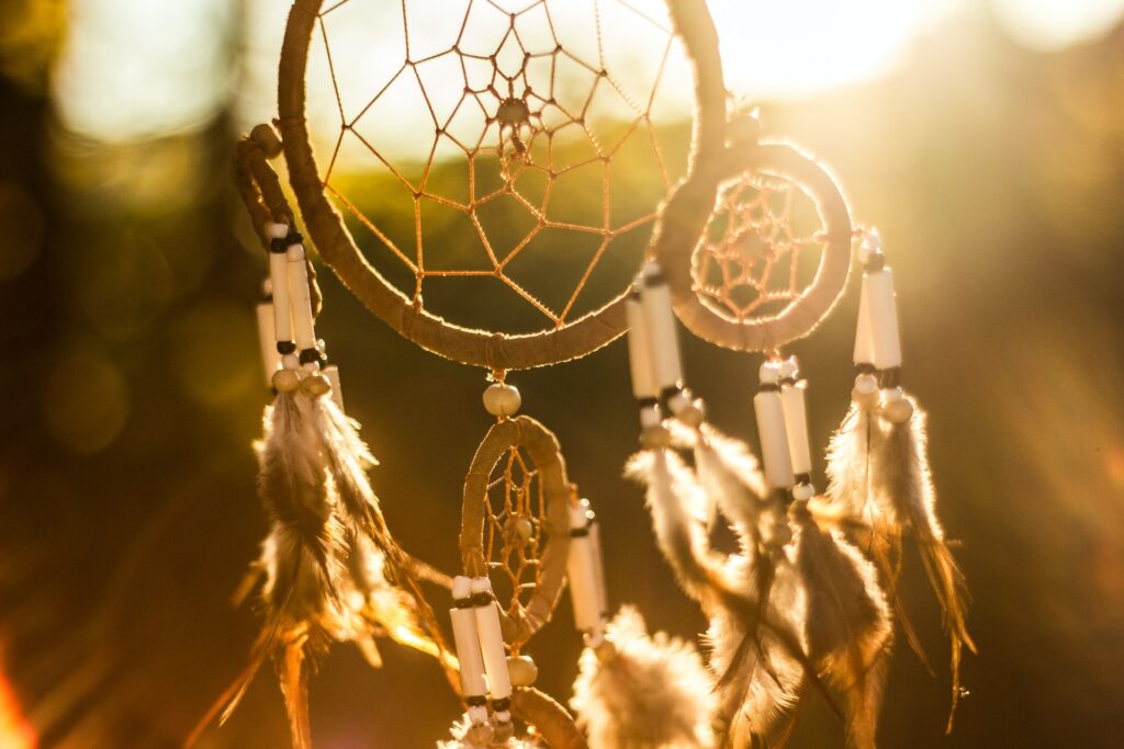 A warm, sunlit close-up shot of a traditional dreamcatcher hanging outdoors. The sunlight streams in from behind, creating a beautiful, bright golden glow and a soft bokeh effect in the background. The dreamcatcher features a circular wooden web woven with string, decorated with dangling white beads and soft feathers that catch the bright light.