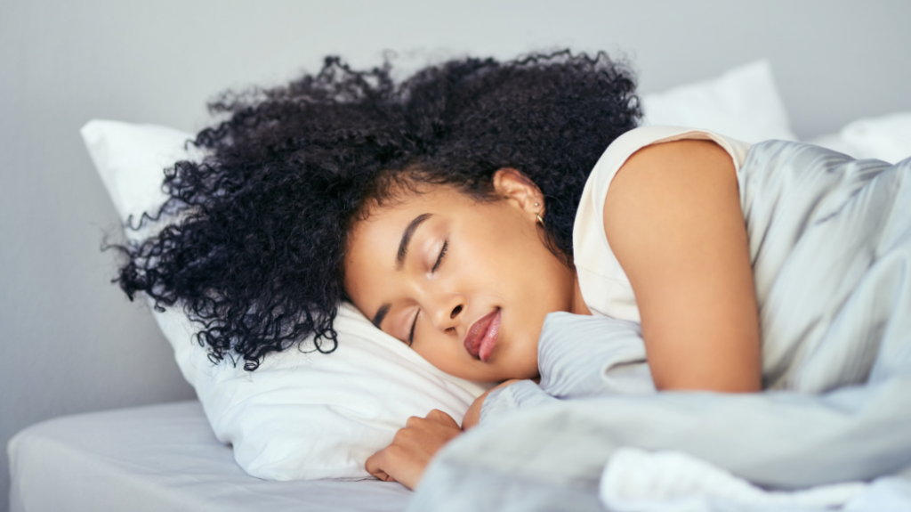 A close-up, serene photograph of a woman with voluminous, dark curly hair sleeping peacefully on her side. Her head is resting on a fluffy white pillow, and she is tucked comfortably under a light gray duvet, showing a calm and relaxed facial expression.